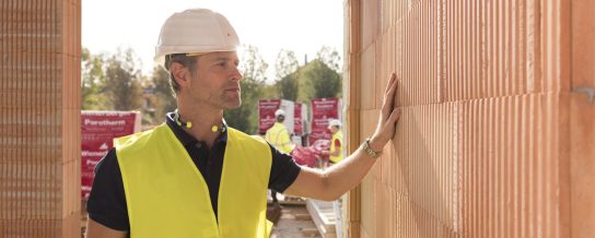 Builder on construction site touching clay block wall, construction workers and clay block pallets in the background