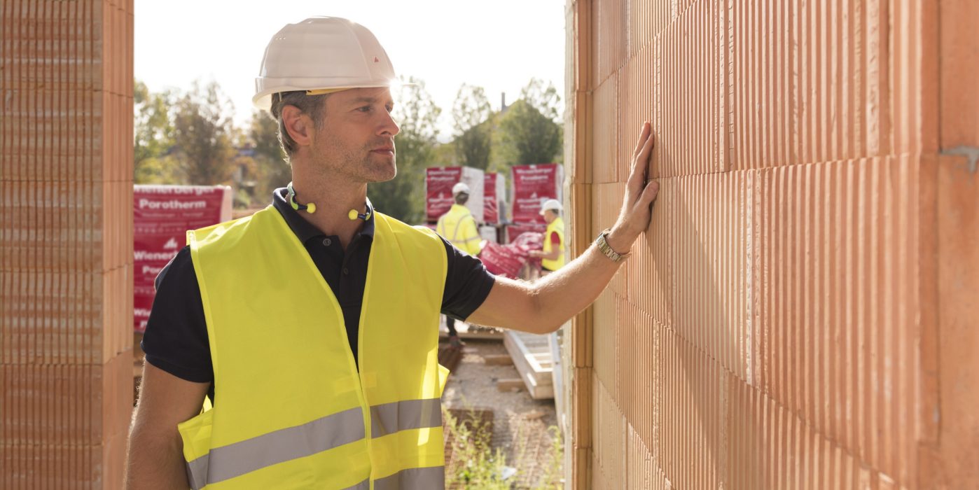 Builder on construction site touching clay block wall, construction workers and clay block pallets in the background