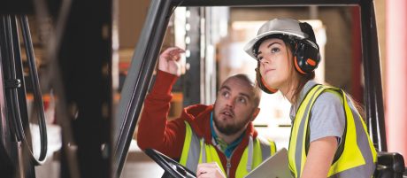 A young female forklift driver is being trained on approaching the racking of the warehouse . A male colleague or trainer can be seen advising her on how to perform the manoeuvre . She is wearing a hi vis jacket , hard hat and ear defenders .